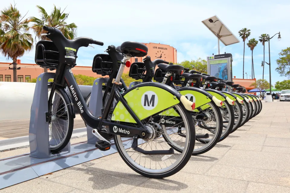 Row of black and green Los Angeles Metro bike share bikes at station with red building with clock and'Los Angeles' lettering in background.